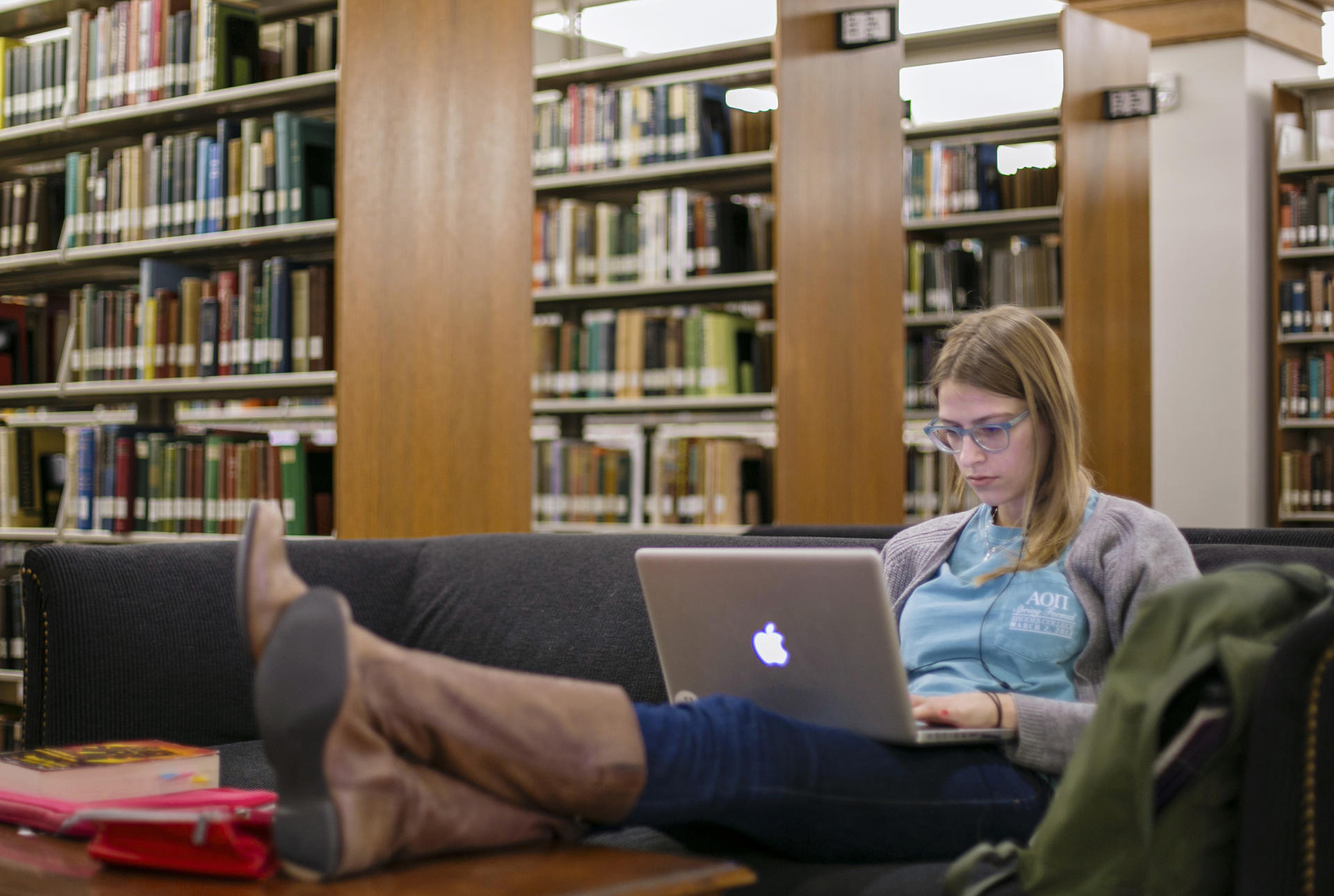 A female student sits with her feet propped up on the table in the library, staring at her laptop screen.