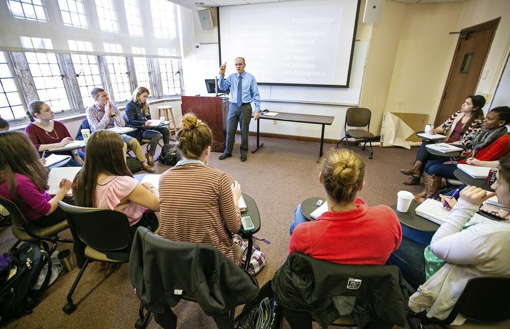 A professor stands at a whiteboard, with large stained glass windows to his right, while a class sits in the round, watching in rapt attention.