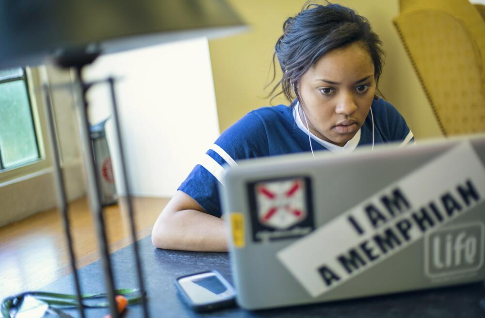 A student at a library table looks at a laptop with a "I AM A MEMPHIAN" sticker on the back.