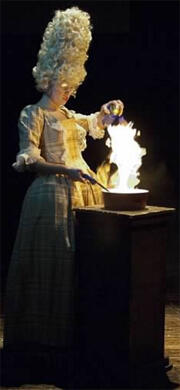 A woman in an 18th century dress and wig makes fire in a pan on a pedestal