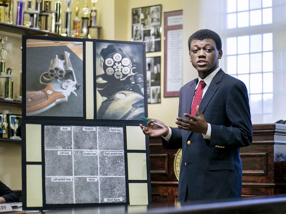 A young man in a suit and red tie points to exhibits arrayed in a practice courtroom.