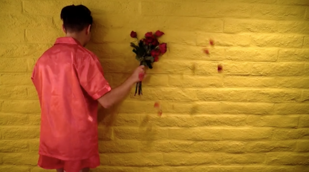 a  figure in a red shirt faces a yellow wall and holds a bouquet against the wall