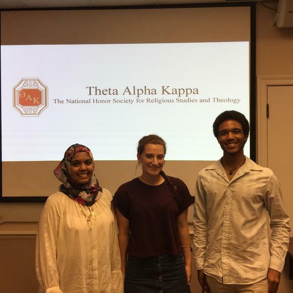 three young people in front of a sign for Theta Alpha Kappa