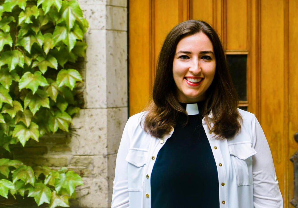 A young, dark haired woman in a clerical vestments smiles warmly.