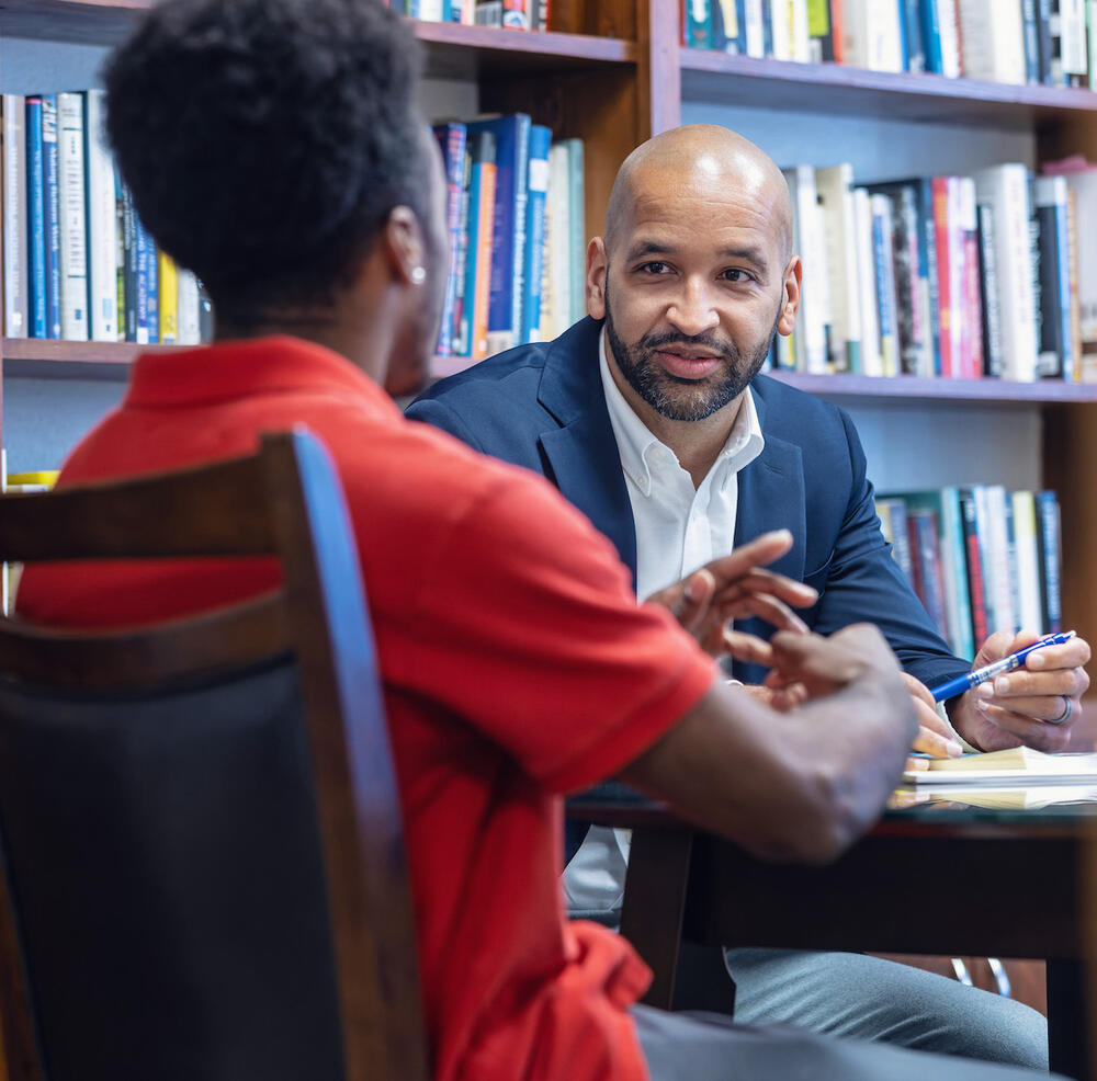 A man talking emphatically to a student seated across the table from him.