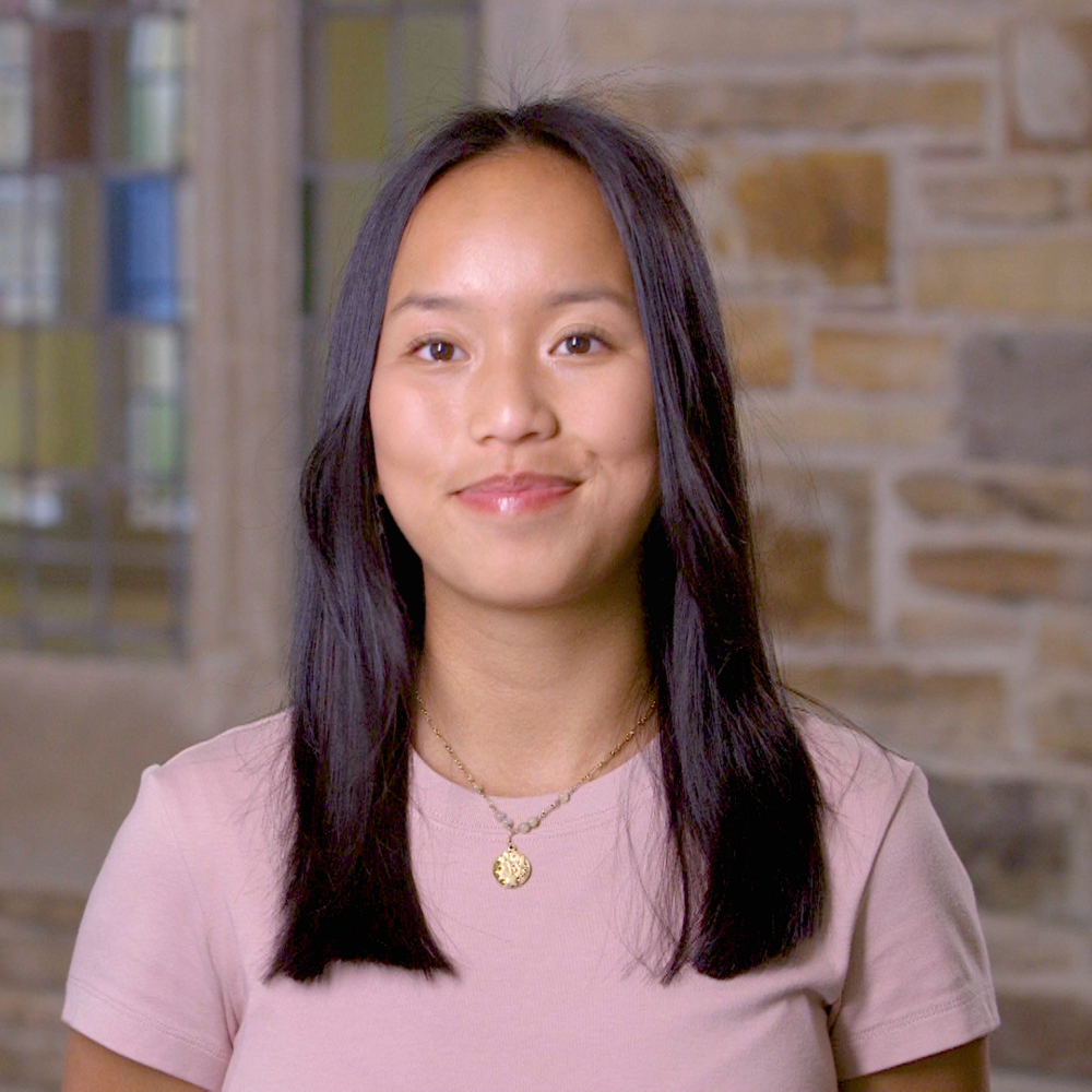 A young woman with straight black hair in a short sleeved pink v-neck.