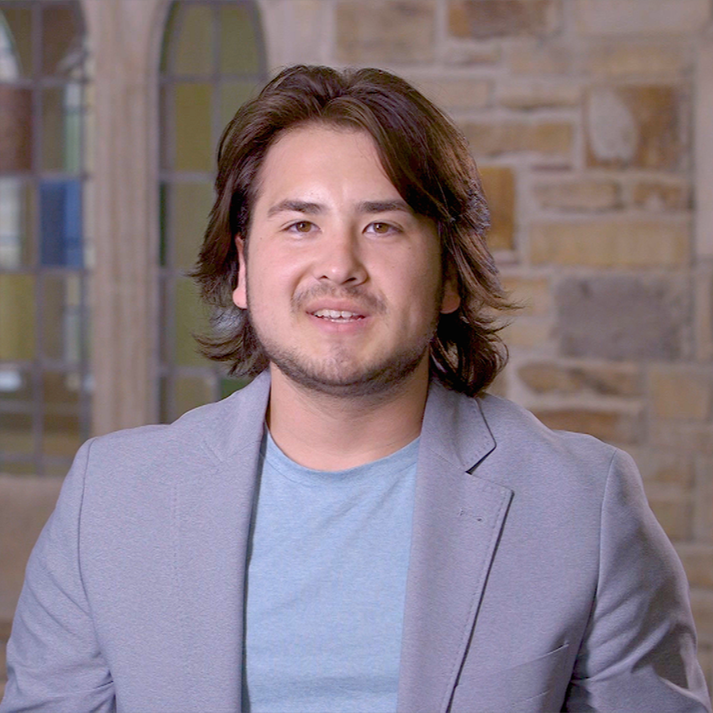 A young man with long brown hair, a light blue ring neck shirt and light gray sports jacket.