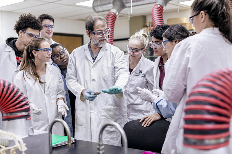 a professor shows a group of students in lab coats an experiment