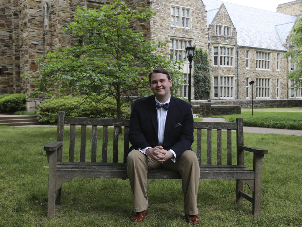 a young man in a suit sits on a wooden bench in front of a Gothic building