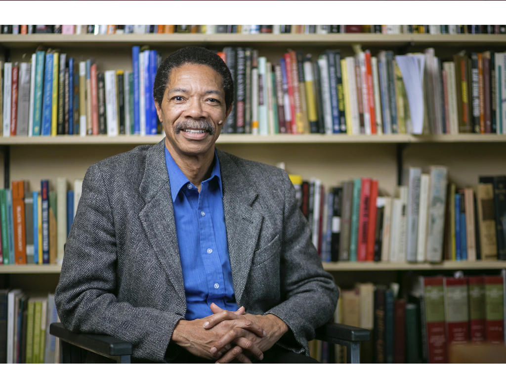 an african american male professor sitting in a chair in front of a filled bookshelf